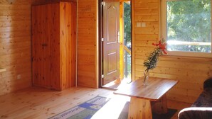 A wooden cabin interior with a wooden door, a window with a view of trees, a small table with a vase, and a patterned rug. - Wooden House in Lubin by Lake Wicko (Międzyzdroje)