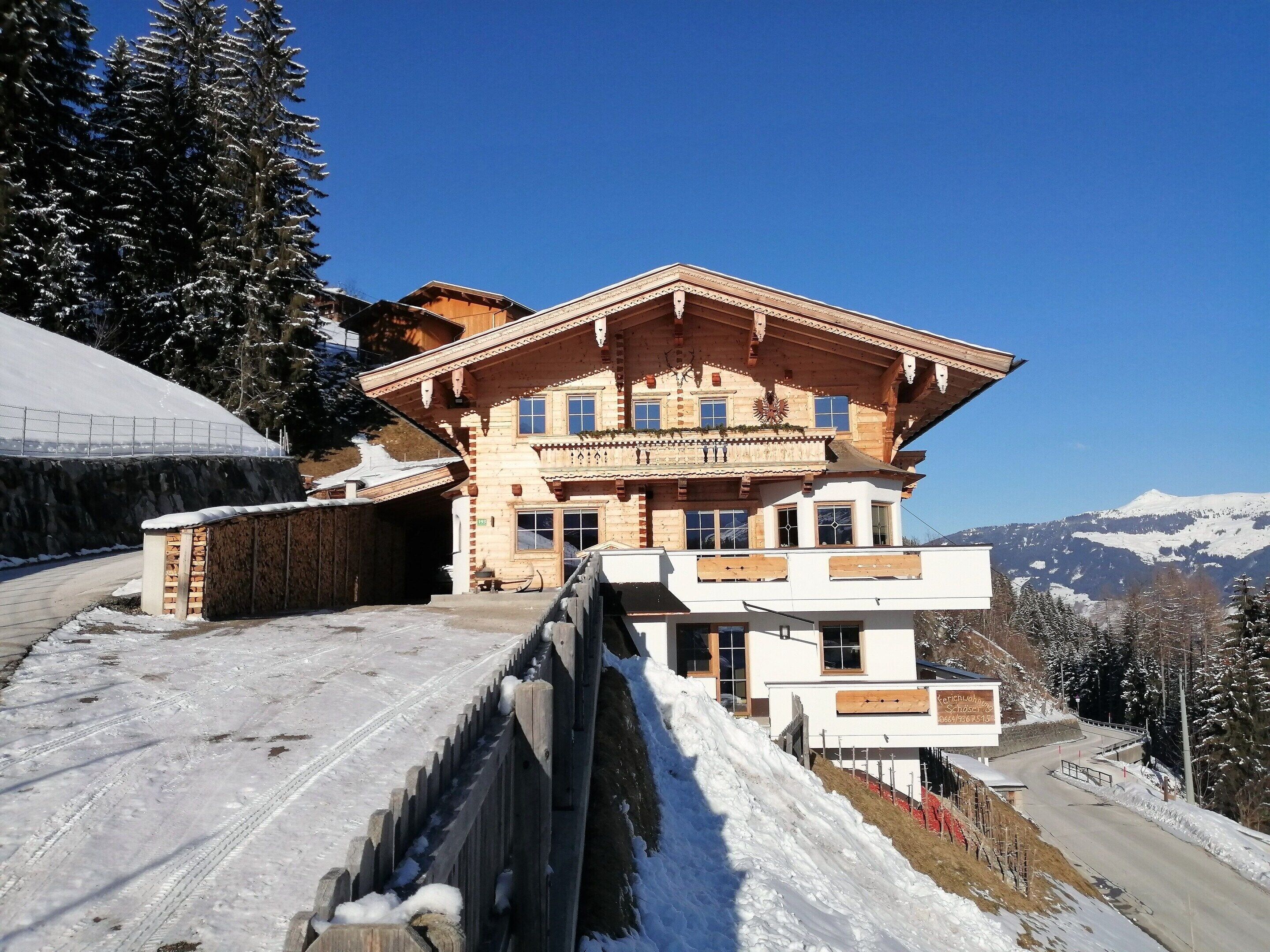A large wooden chalet with a balcony, surrounded by snow-covered trees and mountains in the background.