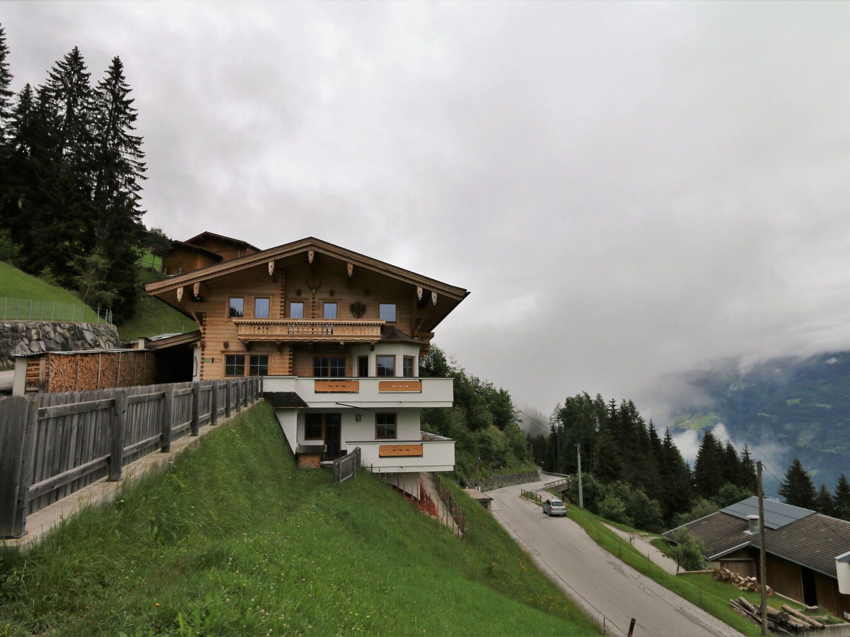 A two-story chalet with a balcony, situated on a hillside with a road and forested background.