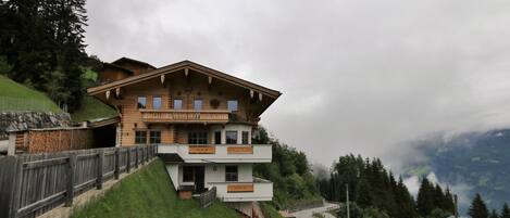 A two-story chalet with a balcony, situated on a hillside with a road and forested background.