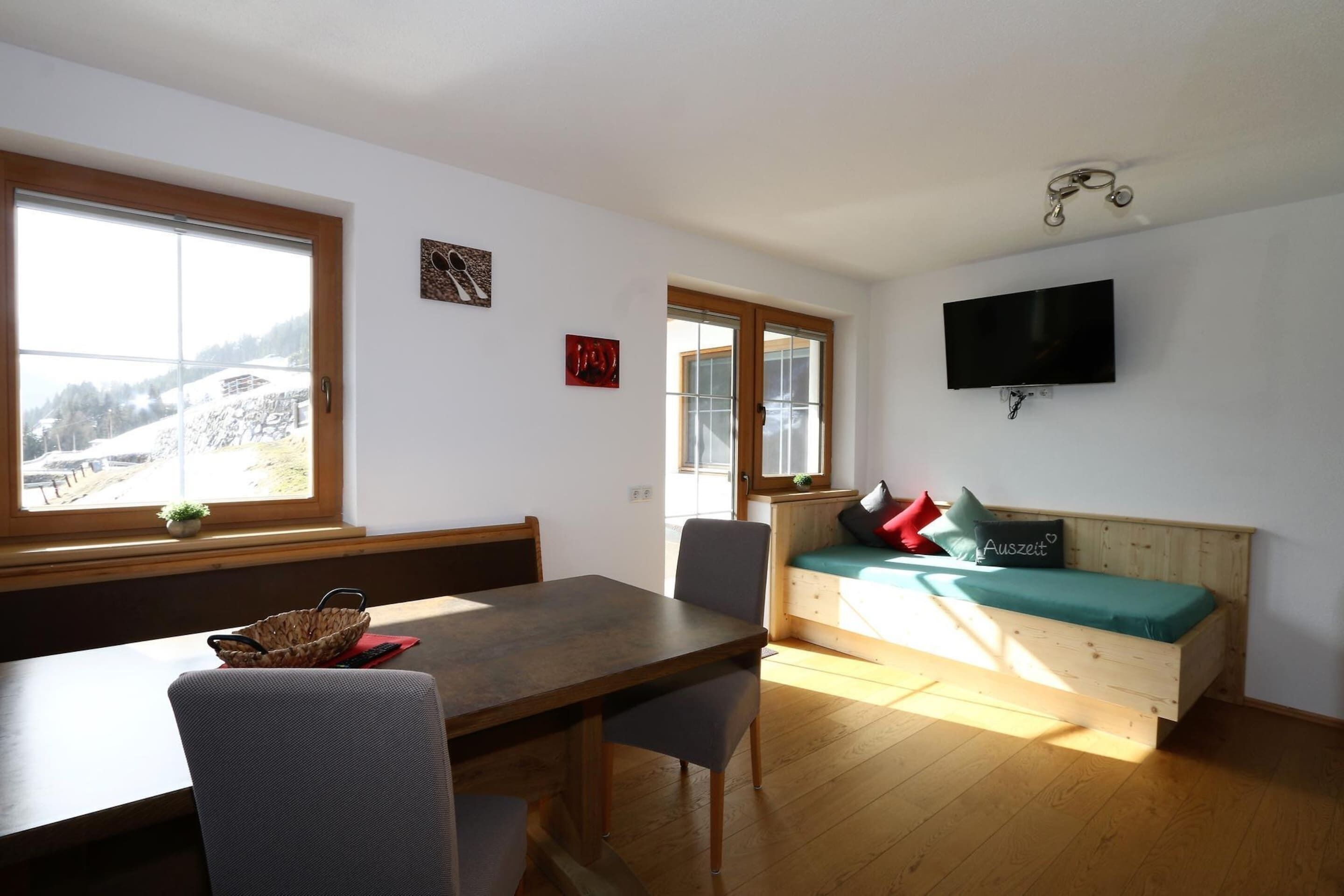 Een kamer met een houten eettafel, een bank met kussens, een aan de muur bevestigde televisie, en een raam met uitzicht op een besneeerd landschap.