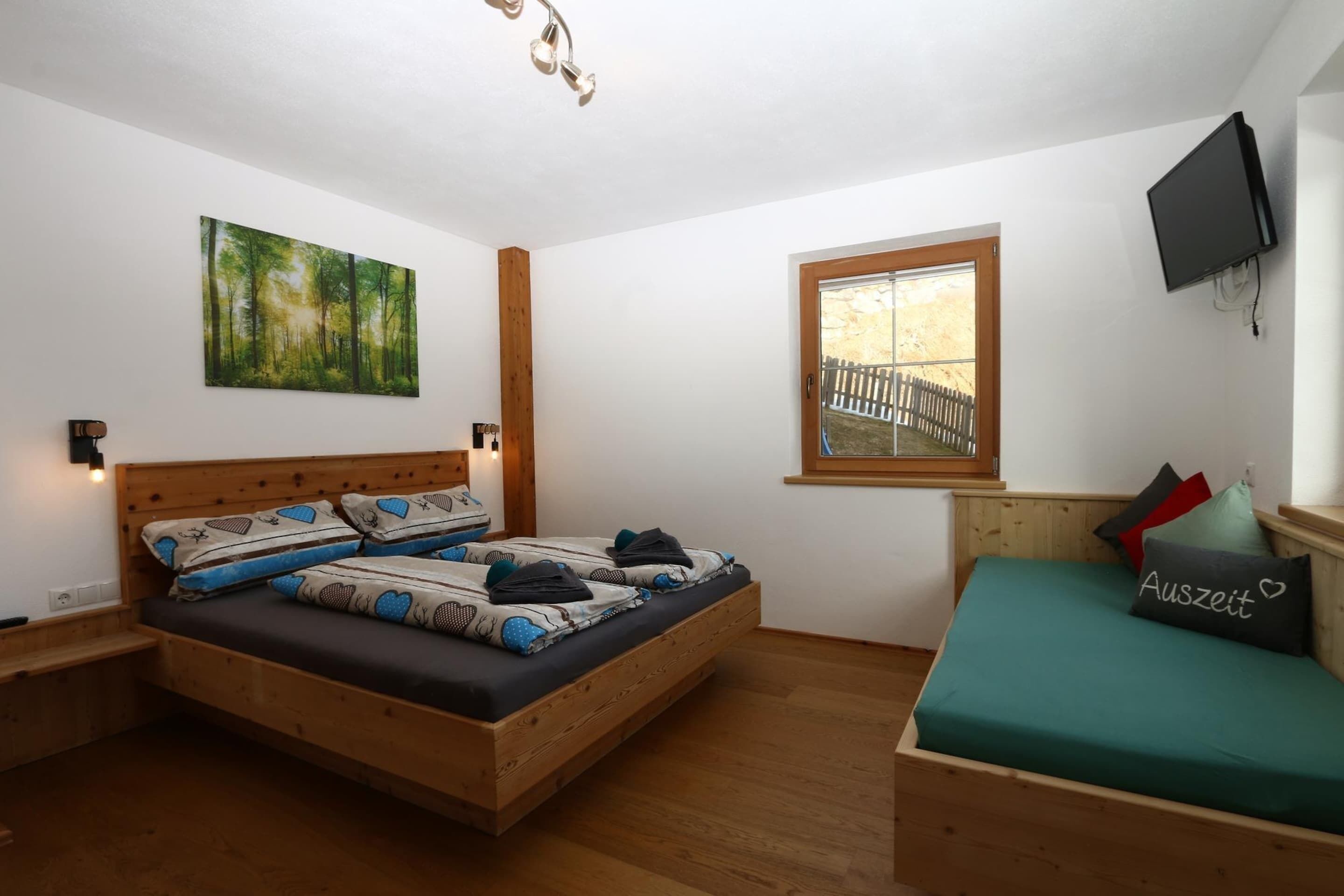 A bedroom with a wooden bed, a window with a view of a wooden deck, a painting of a forest, and a mounted TV.