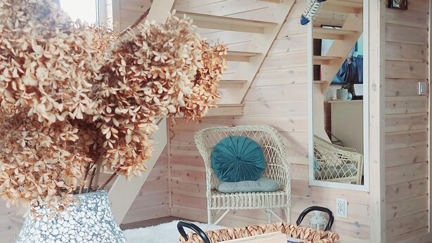 A cozy room with a wooden ceiling, a wicker chair with a blue cushion, a woven basket on the floor, and a vase with dried flowers. - Blue Lagoon - Vacation Home in Nowe Warpno (Nowe Warpno)