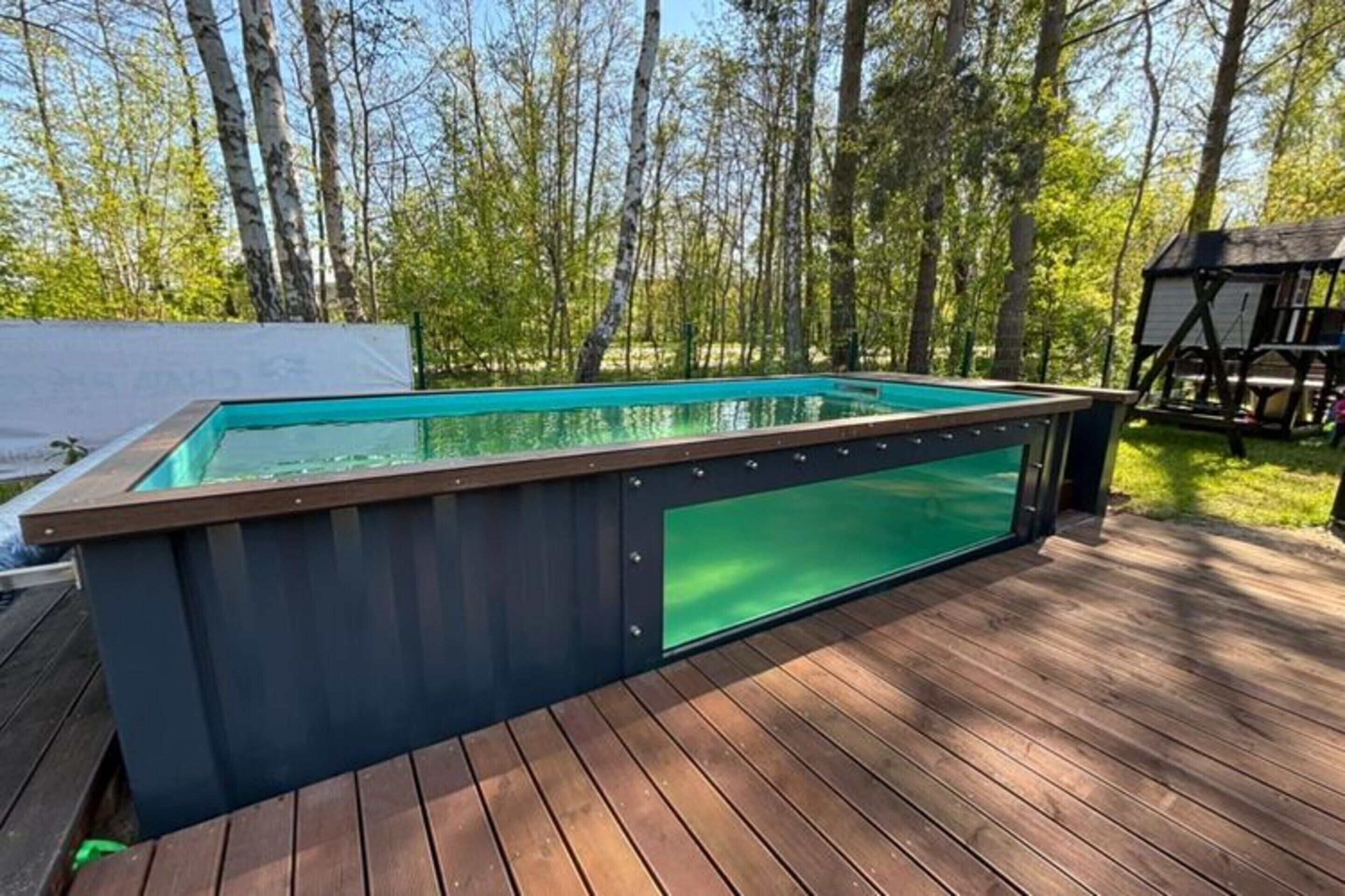 Ein rechteckiger Pool mit dunklem Sonnendeck und Glasboden, umgeben von Holzdeck und Bäumen.