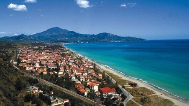 Luftaufnahme einer Küstenstadt mit Strand, Häusern und Bergen im Hintergrund.