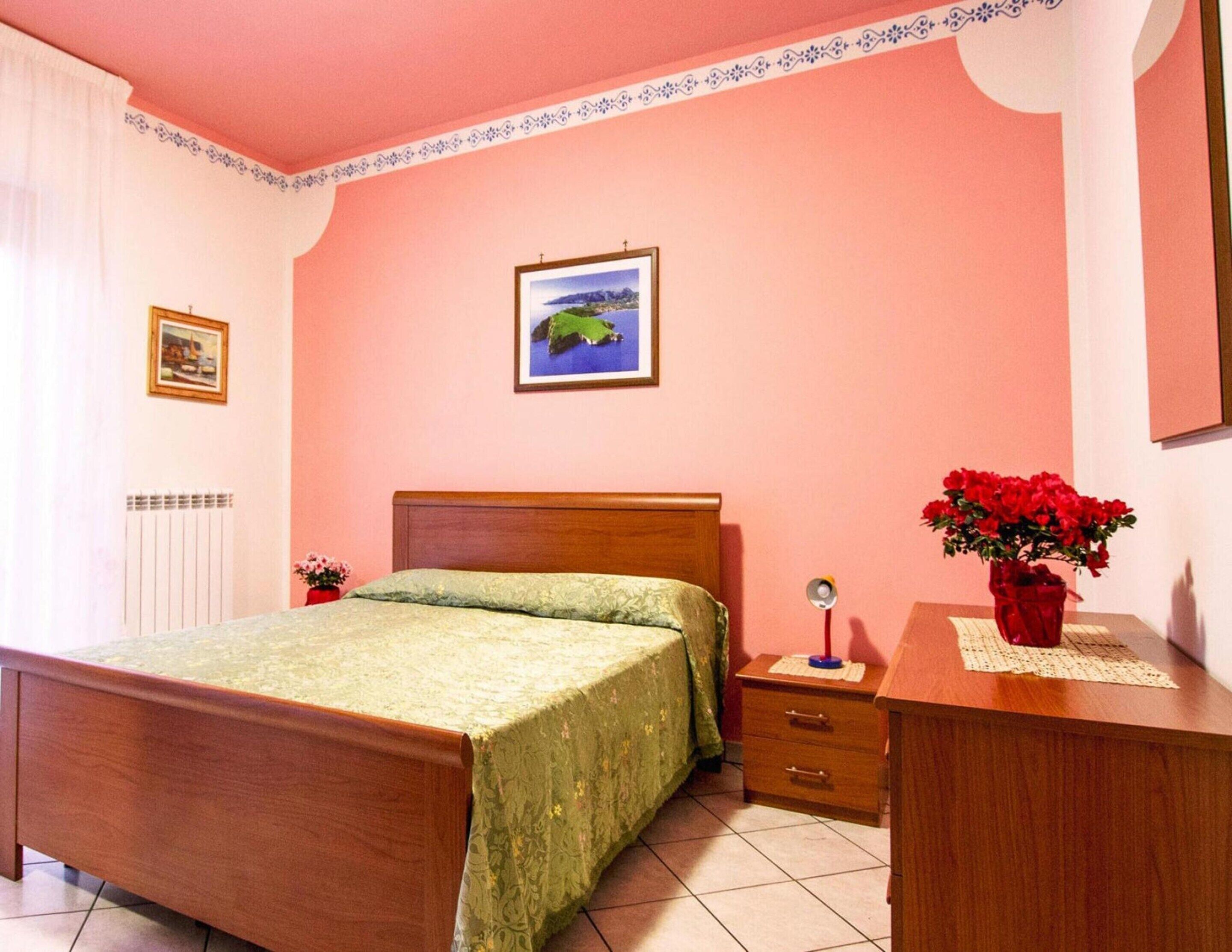 Un dormitorio con cama de madera, una mesita de noche, un aparador, un cuadro enmarcado en la pared y un jarrón con flores rojas.