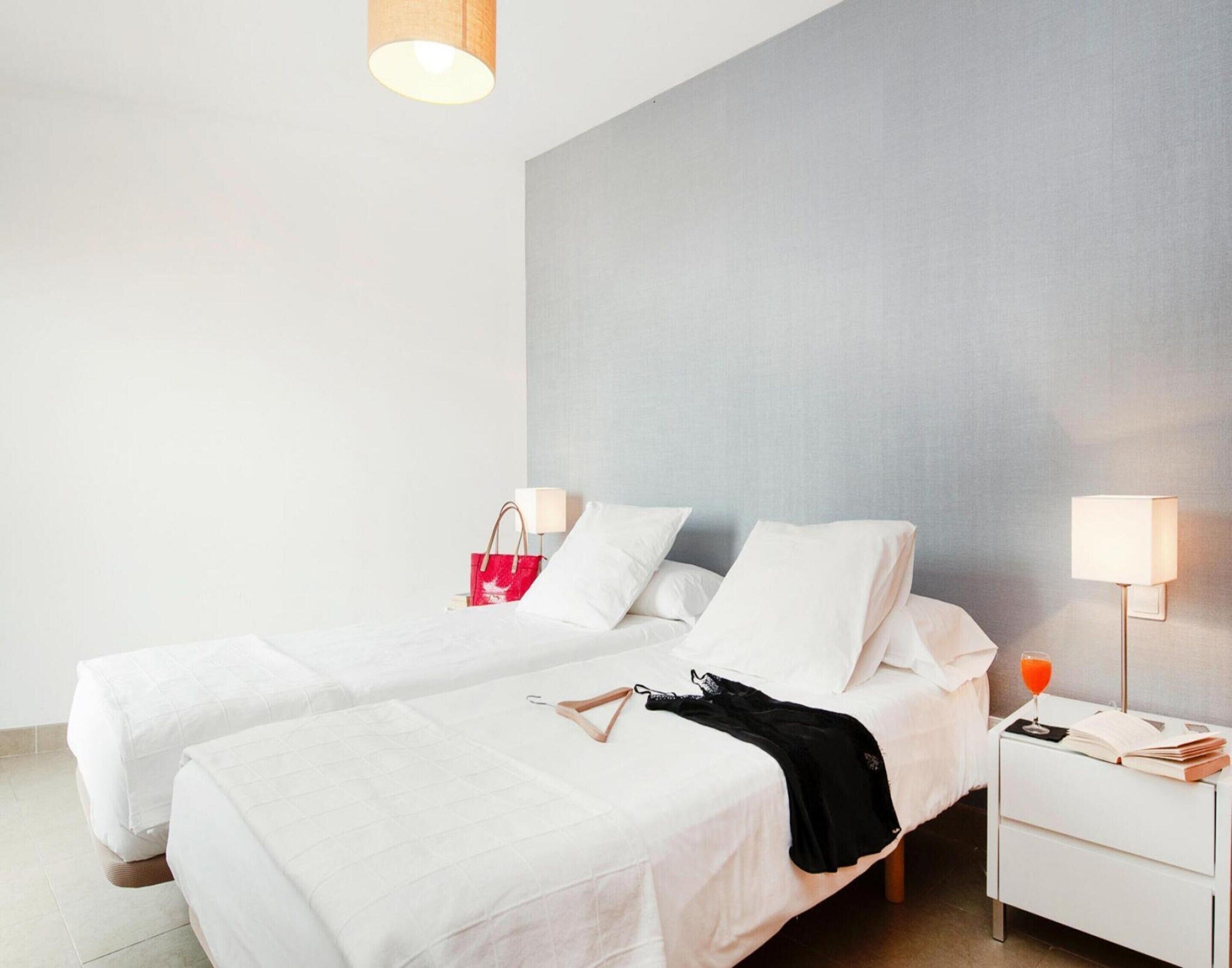 Una habitación de hotel moderna con dos camas individuales, una mesita de noche con lámpara y una mesita auxiliar, y un bolso rojo a la izquierda.