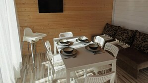 A wooden cabin interior with a dining table set for four, white plastic chairs, a sofa with patterned cushions, and a mounted television. - Holiday Homes With Terraces, Close to the Beach (Ustronie Morskie)