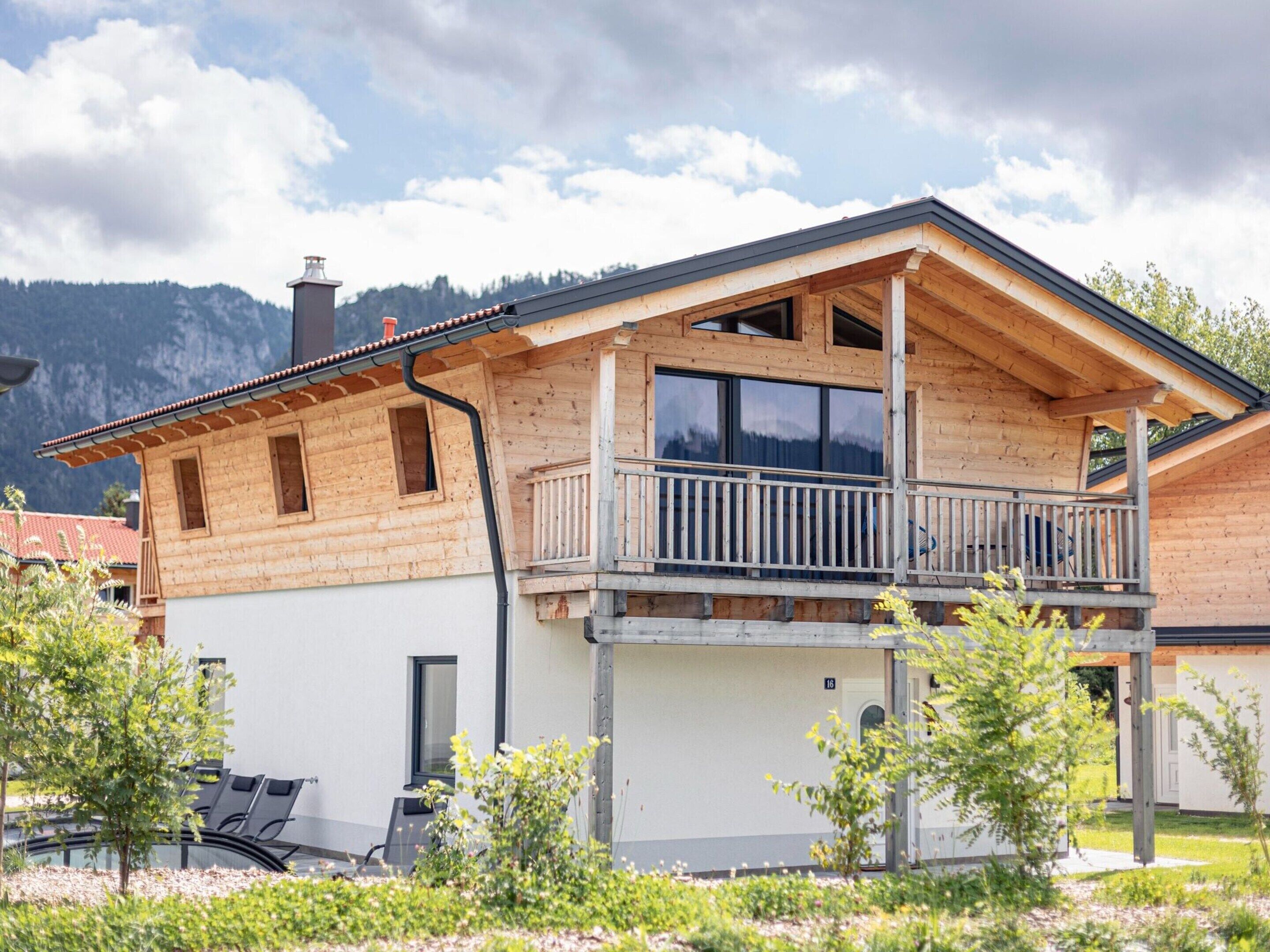 A two-story wooden house with a balcony, large windows, and a view of mountains.