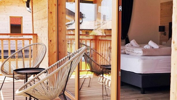 A wooden deck with wicker chairs, a bed with a dark headboard, and a view of a building with a red roof through a glass door. - Inzell Chalets With Private Pool (Inzell)