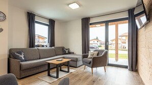 A modern living room with a sectional sofa, a small table, and a chair. There is a large window with curtains and a view of the outside. - Inzell Chalets With Private Pool (Inzell)