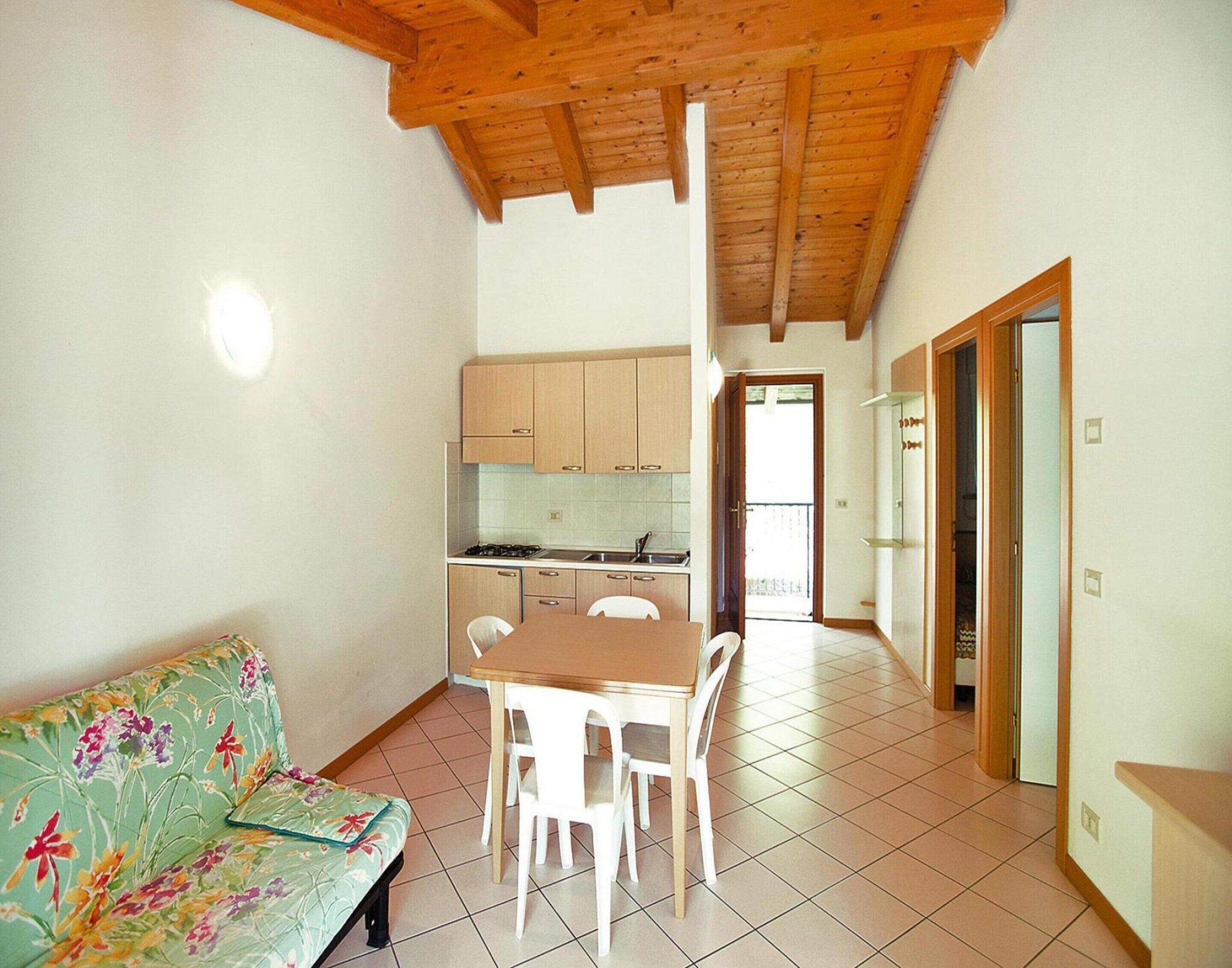 A small, open-plan living space with a wooden ceiling, a kitchenette, a dining area with a table and chairs, and a sofa with a floral cushion.