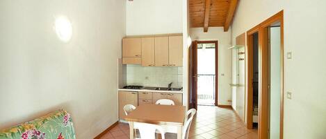 A small, open-plan living space with a wooden ceiling, a kitchenette, a dining area with a table and chairs, and a sofa with a floral cushion.