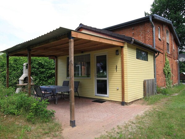 A small, yellow wooden house with a brick exterior, a covered patio with a table and chairs, and a chimney. - Ferienhaus in Petersdorf mit Schonem Garten (Fuenfseen)