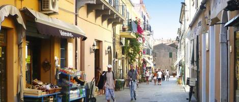 A narrow street with shops and balconies, stone pavement, and pedestrians.