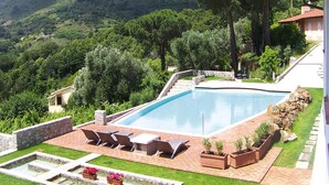 A swimming pool with a patio area, surrounded by a stone wall and lush greenery, overlooking a hilly landscape. - Apartment in Elba With Panoramic Views (Rio)