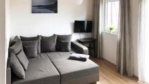 A modern living room with a grey sectional sofa, a flat-screen TV, a small table, and a window with curtains. - Ferienhaus am Partwitzer See, Elsterheide (Elsterheide)