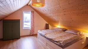 A bedroom with a sloped ceiling, a bed with a patterned bedspread, a green wardrobe, and a window with floral curtains. - Ferienhaus am Partwitzer See, Elsterheide (Elsterheide)