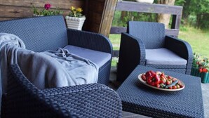 Un conjunto de sofá y sillones de mimbre en un porche con barandilla de madera, una mesita con una fuente de fruta, y plantas en macetas al fondo.