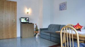 Olohuoneessa on harmaa sohva, puinen ruokapöytä, puinen kaappi, televisio ja pieni kasvi.