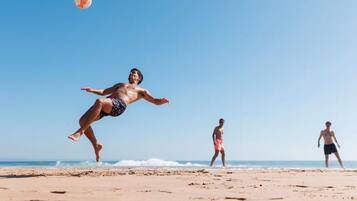 Escena de playa con una persona saltando y un disco volador en el aire, y otras dos personas caminando al fondo.