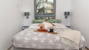 A neatly made bed with a floral duvet, a bedside table with a lamp, and a window with a view of buildings. - Holiday Apartment Right on the Beach, Pobierowo (Rewal)