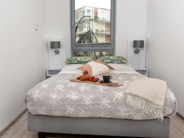 Un lit bien fait, recouvert d’une couette à motifs floraux, une table de chevet avec une lampe et une fenêtre donnant sur des immeubles.