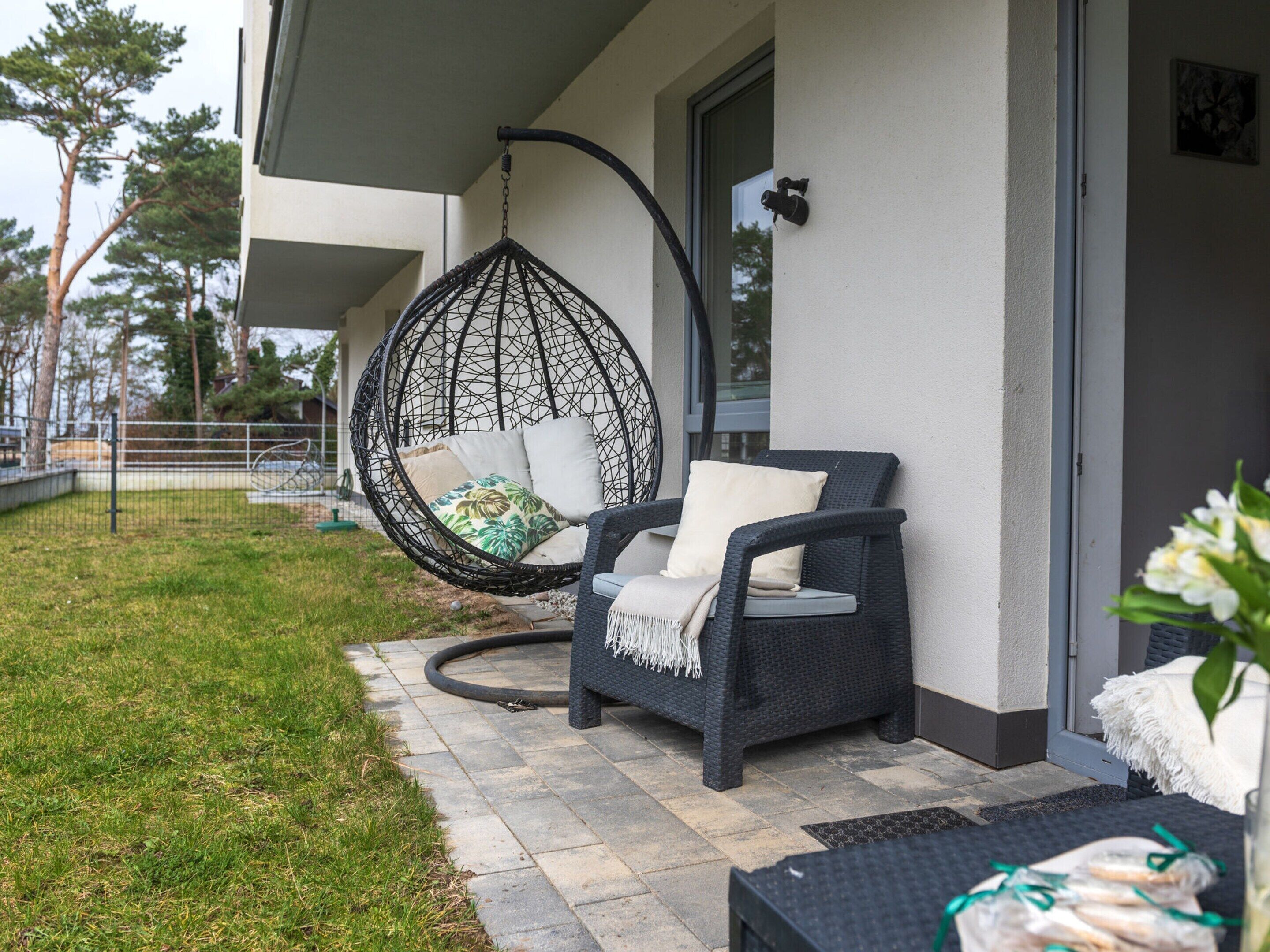 Ein Korbstuhl mit Kissen und ein Hängesessel auf einer Terrasse mit einem gepflasterten Weg.