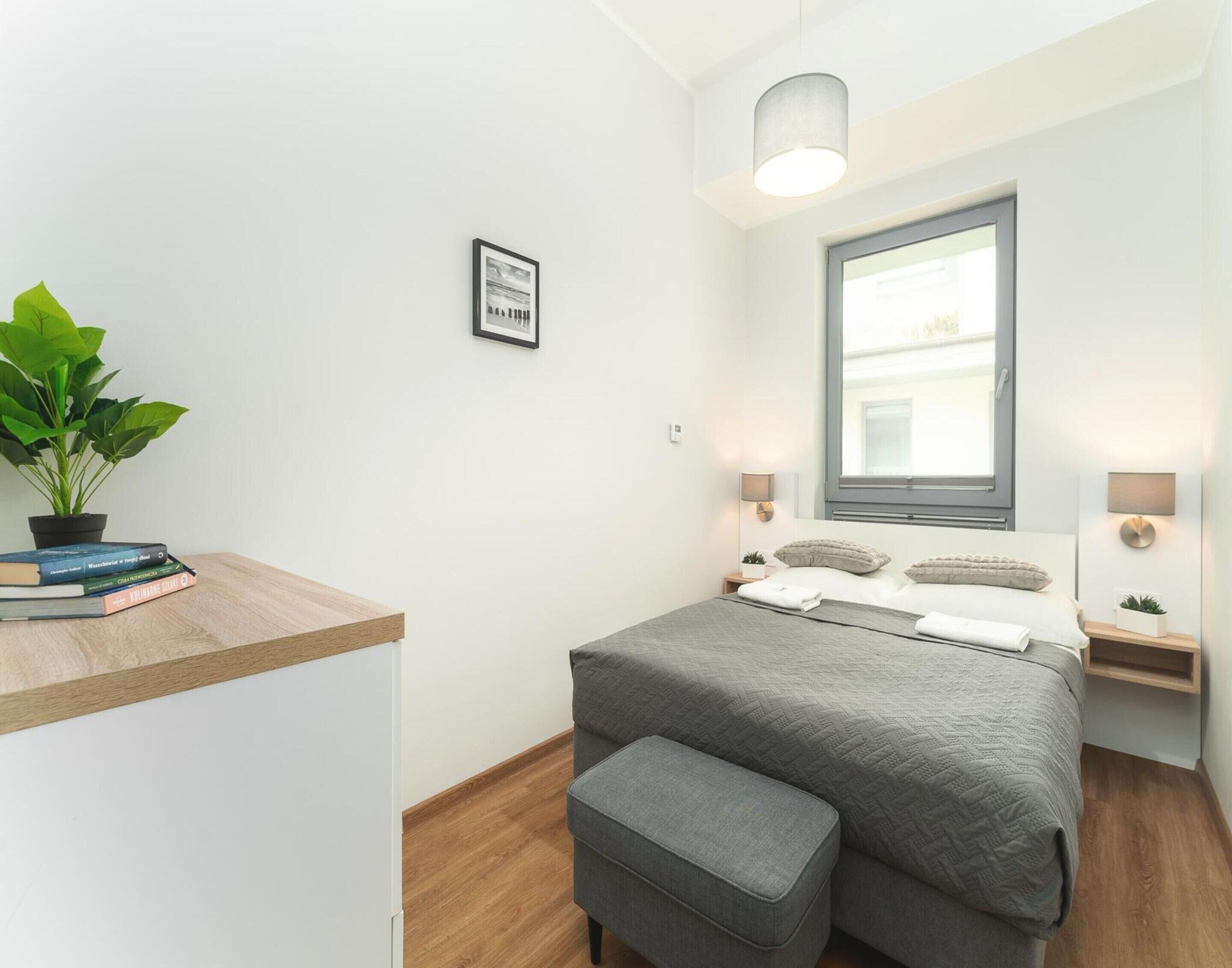 A modern bedroom with a bed, bedside tables, a window, a potted plant, and a small desk with books.