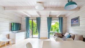 En moderne stue med et fladskærms-tv, hvide møbler, et spisebord og udsigt til grønt område gennem store vinduer.