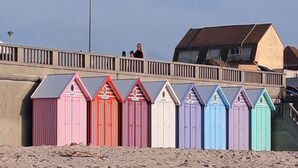 A row of colorful beach huts along a sandy beach, with a bridge overhead. - Ferienwohnungen, Wyk auf Fohr (Wyk auf Foehr)