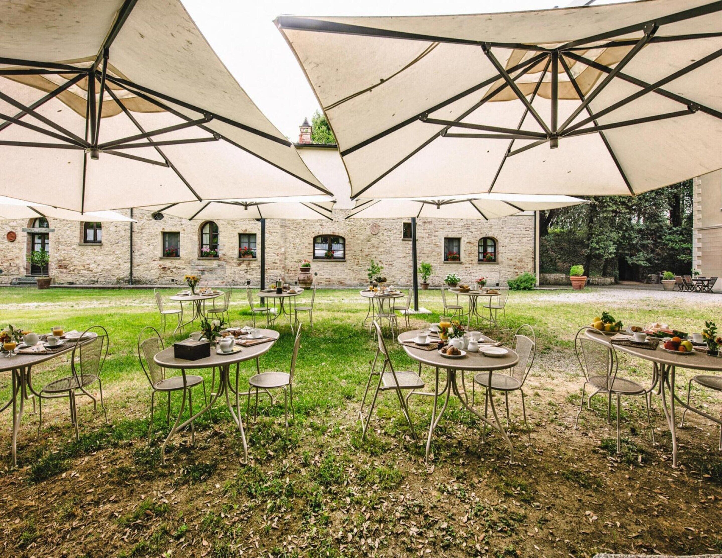 Un área de comedor al aire libre con mesas y sillas bajo sombrillas grandes, frente a un edificio de piedra con ventanas y un jardín.