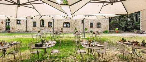 Un área de comedor al aire libre con mesas y sillas bajo sombrillas grandes, frente a un edificio de piedra con ventanas y un jardín.