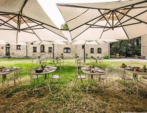 An outdoor dining area with tables and chairs under large umbrellas, set against a stone building with windows and a garden. - Apartment in Santo Pietro Belvedere With Pool-formerly TUI Ferienhaus (Capannoli)