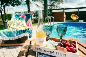 Ein Poolbereich mit einem Holztisch, auf dem eine Flasche, Gläser und eine Kiste mit frischem Obst stehen.