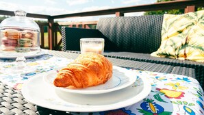 A croissant on a white plate with a floral tablecloth, a glass of milk, and a glass container of macarons on a wicker table outdoors. - Holiday Home With Private Sauna, Jacuzzi and Pool (Koscierzyna)