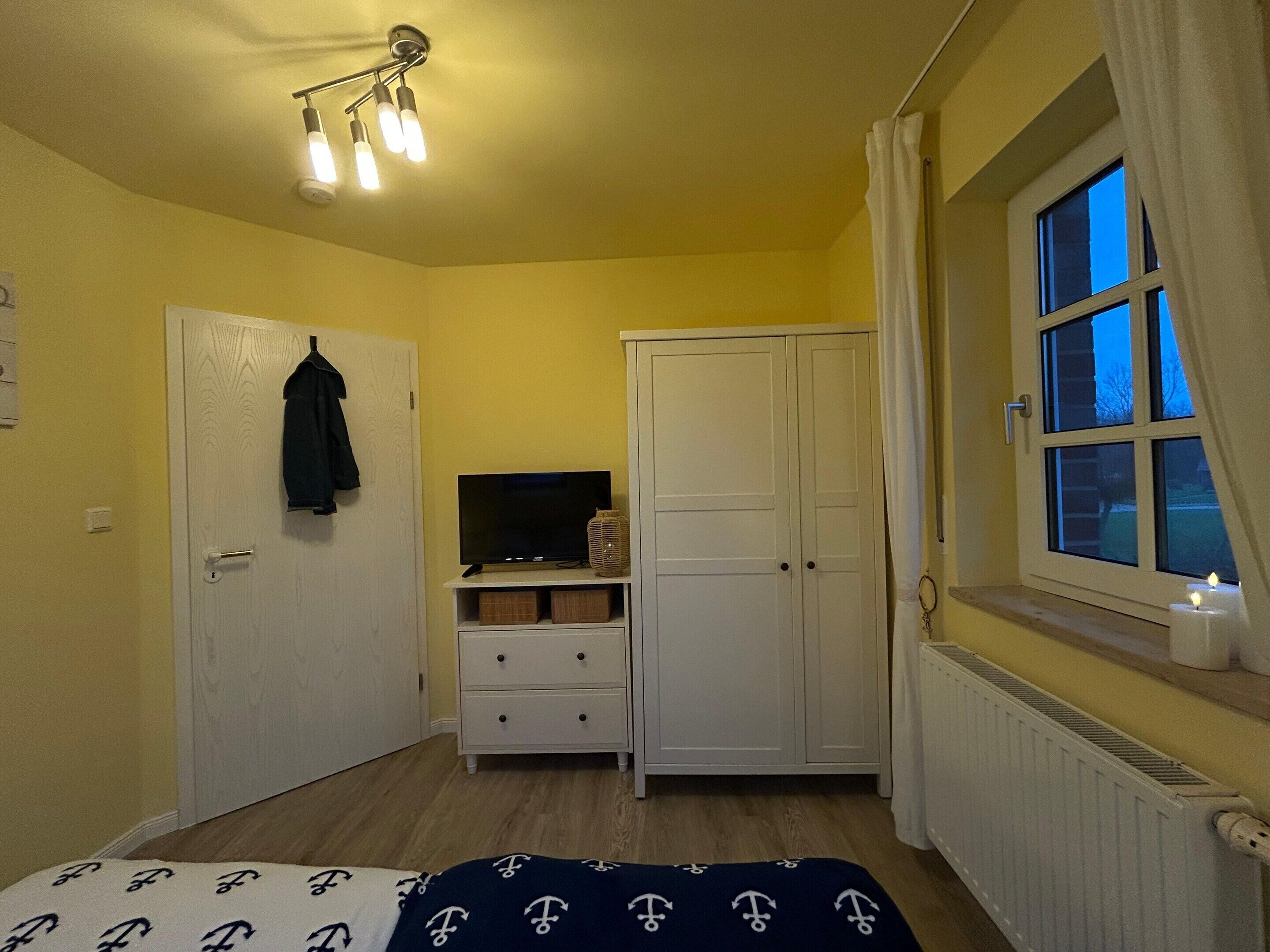 Un dormitorio con cama, televisor sobre un mueble, armario y ventana con cortinas.