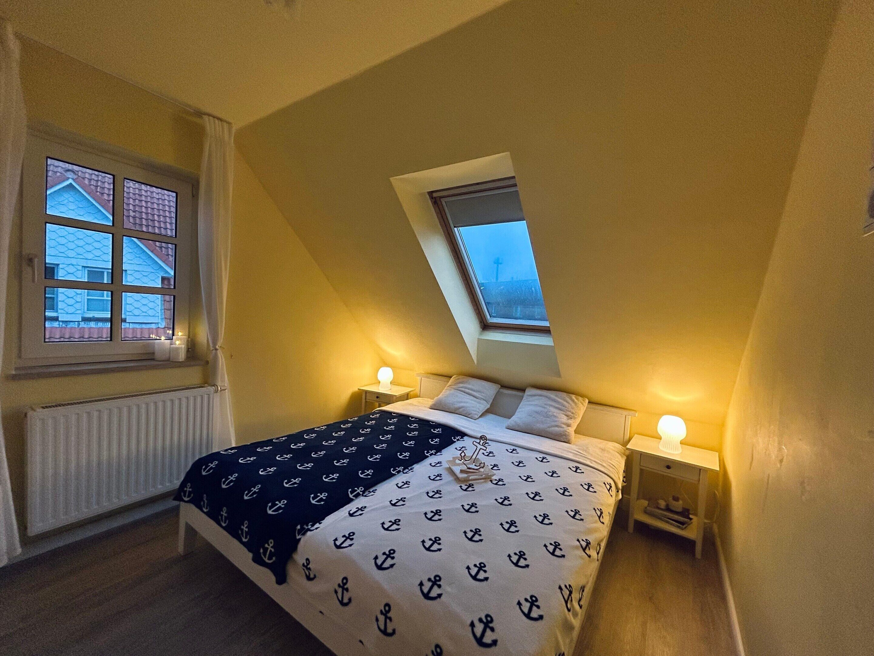 A bedroom with a sloped ceiling, a bed with a patterned bedspread, two bedside lamps, a window with a view of buildings, and a radiator.