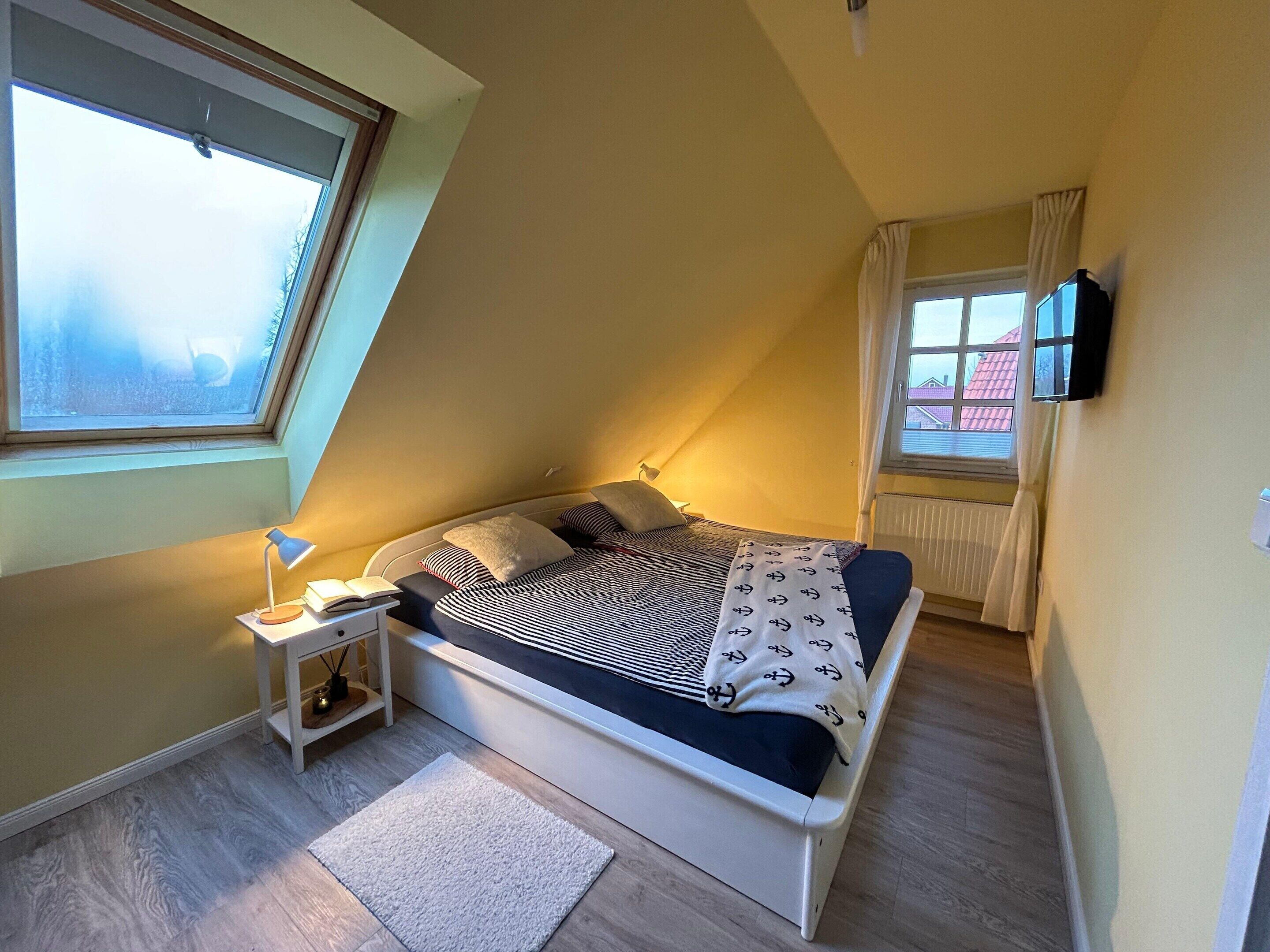 A bedroom with a sloped ceiling, a bed with patterned bedding, a bedside lamp, a small table, and a window with curtains.