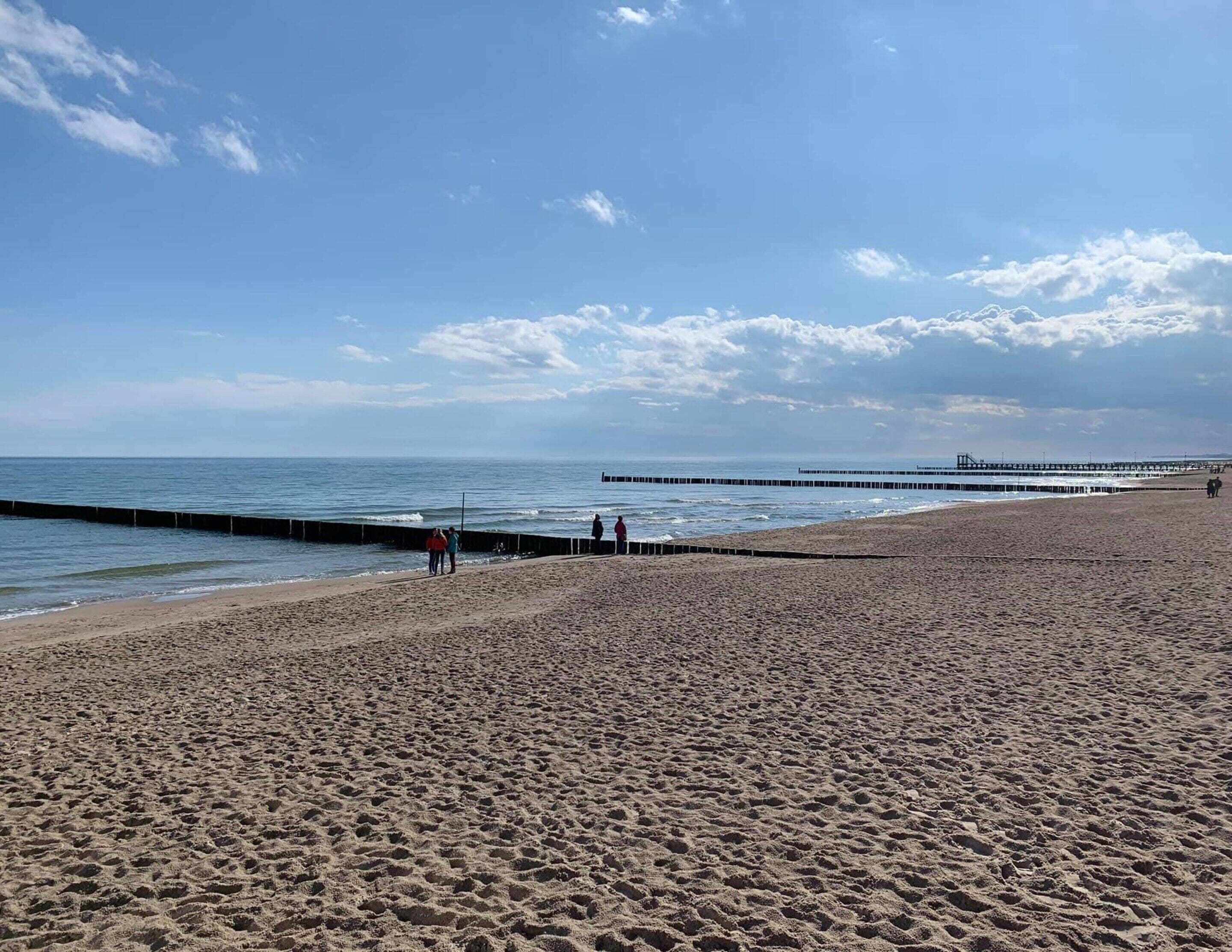 Een breed zandstrand met een kalme zee, een steiger die het water in reikt, en een heldere lucht.