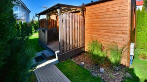 Eine Holzblockhütte mit Veranda und kleiner Terrasse, umgeben von Grün und blauem Himmel.