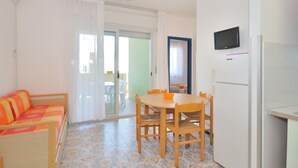 A compact living space with a sofa, a dining table with chairs, a TV mounted on the wall, and a refrigerator. - Apartments Cormoran, Bibione Lido del Sole (San Michele al Tagliamento)