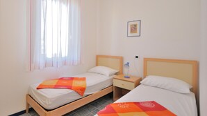 A hotel room with two single beds, a nightstand, a small table, a window with sheer curtains, and a framed picture on the wall. - Apartments Cormoran, Bibione Lido del Sole (San Michele al Tagliamento)