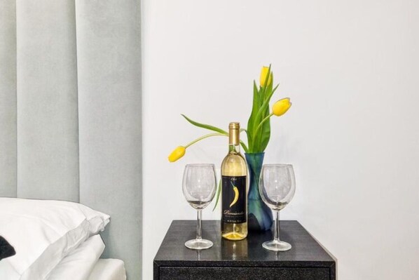 A bed with white linens, a black nightstand with a bottle of wine, two glasses, and a vase with yellow tulips. - Unique Apartment With a Balcony in Darlowko (Darlowo)