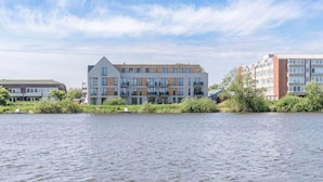 A modern multi-story building with a flat roof, large windows, and a mix of brick and glass facades, located beside a calm body of water. - Appartement in Fantastischer Lage auf Nordstrand (Nordstrand)