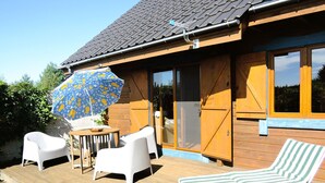 A wooden house with a tiled roof, a covered patio with a table and chairs, and a striped lounge chair. - Holiday Home in Kolczewo for Nature Lovers (gmina Wolin)