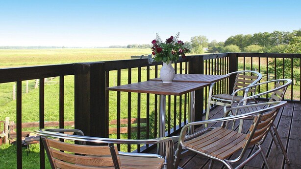 A wooden deck with a table and chairs, a vase with flowers, and a view of a green field. - Apartment Wiesenschein, Moenkebude (Monkebude)