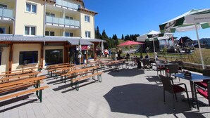 Un espace extérieur aménagé avec des bancs et des tables en bois, un bâtiment à étages et un ciel d’un bleu éclatant.