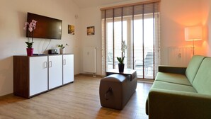 Un salon comprenant un canapé, un téléviseur fixé au mur, un meuble, une table avec une plante et une fenêtre avec des stores.