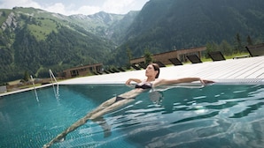 Ein Schwimmbecken mit einer Person im Wasser, umgeben von Bergen und einer Holzterrasse mit Liegestühlen.